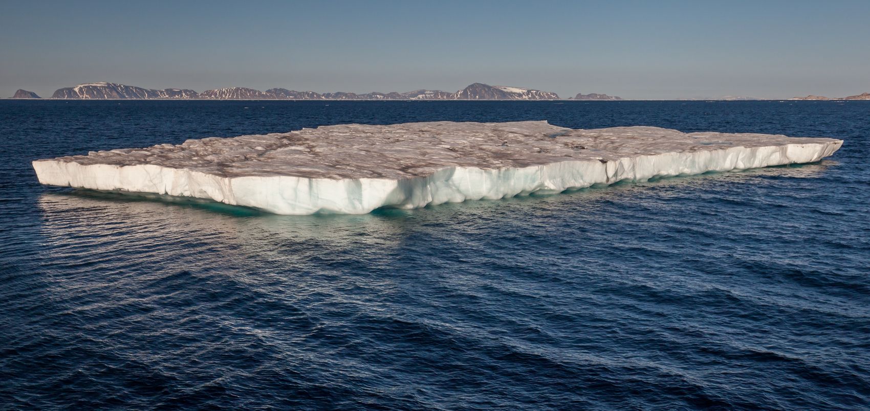 A flat-topped table iceberg floating on dark blue Arctic water west of Sjuøyane, Svalbard, with snow-streaked islands on the horizon