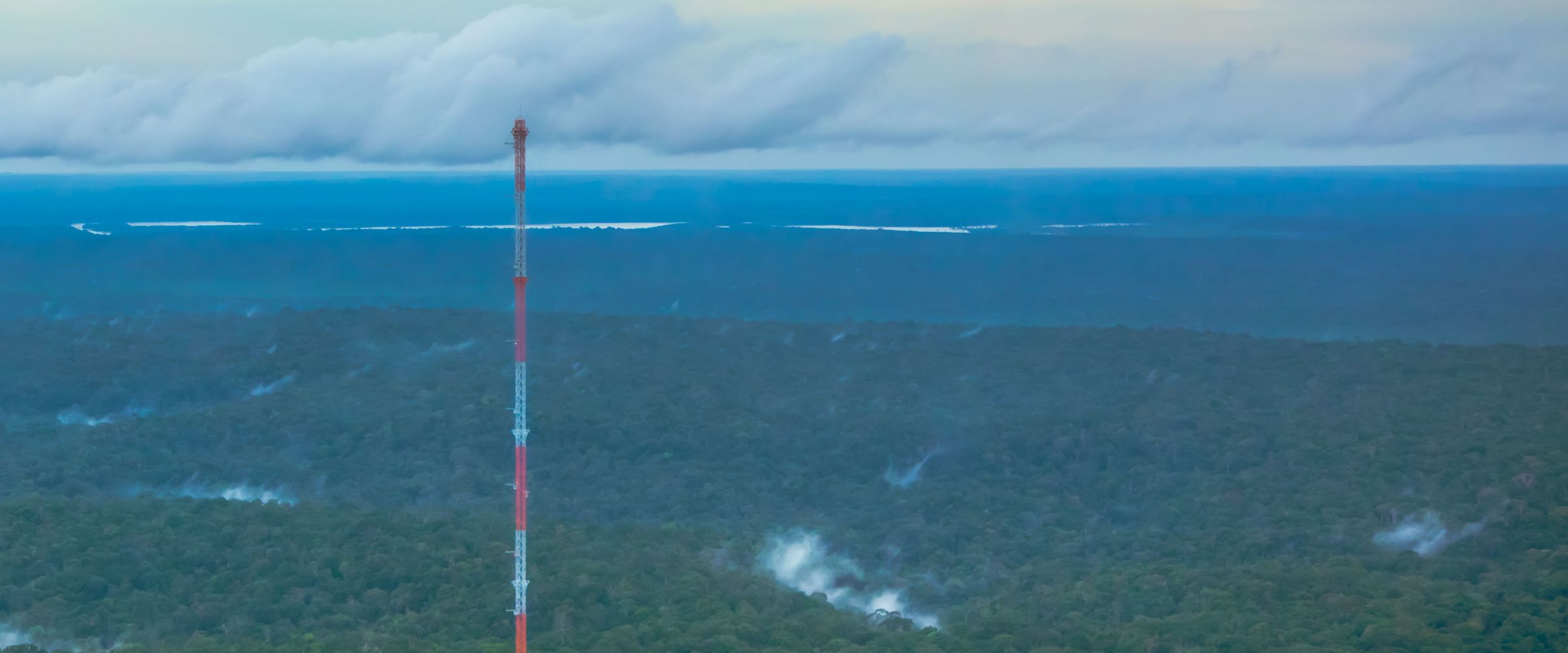 The 325-metre Amazon Tall Tower Observatory rises out of the rainforest canopy, with low mist over the trees and a line of clouds on the horizon