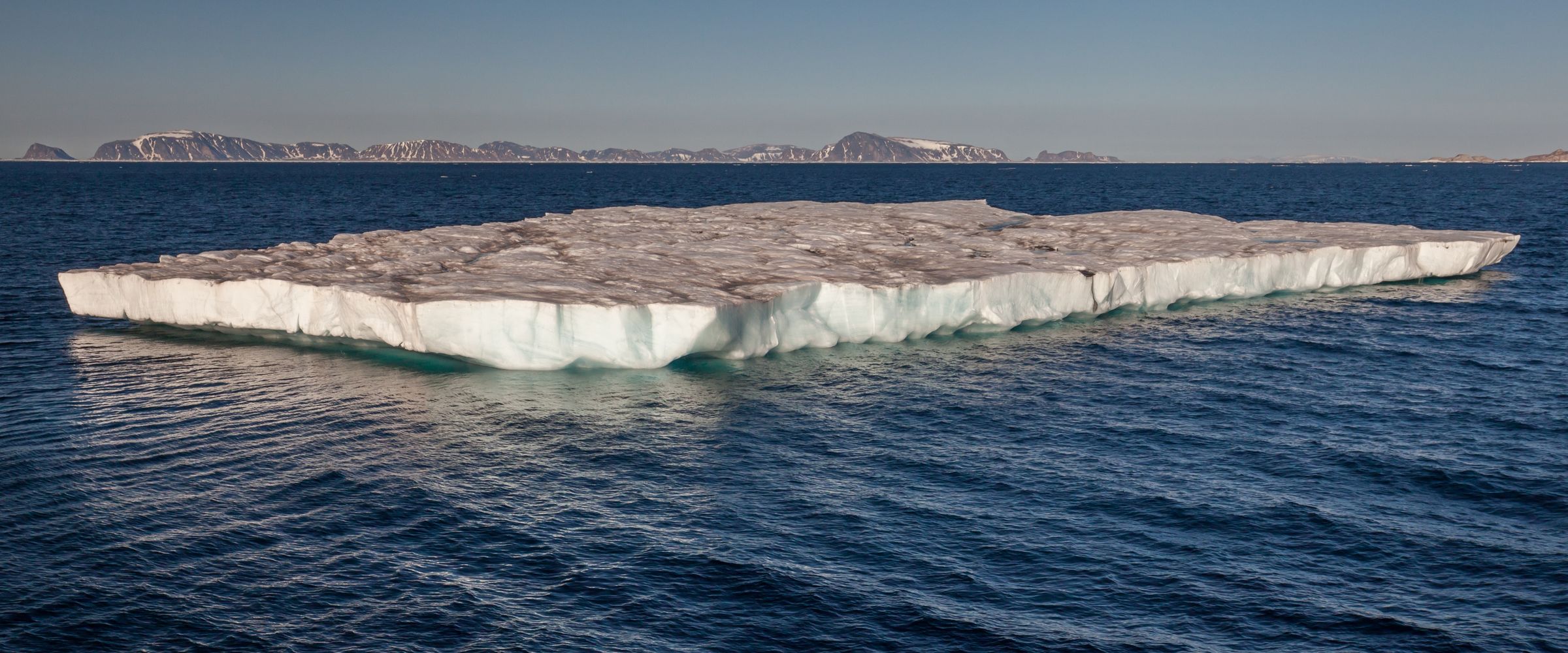 A flat-topped table iceberg floating on dark blue Arctic water west of Sjuøyane, Svalbard, with snow-streaked islands on the horizon